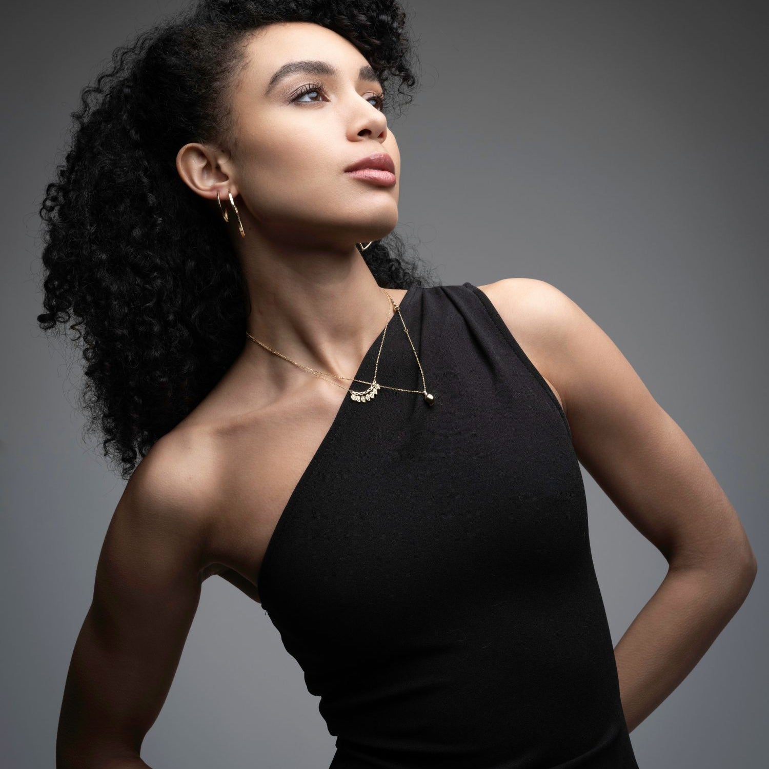 Woman in a black dress wearing gold pendants, one with seven charms and the other a single mango leaf