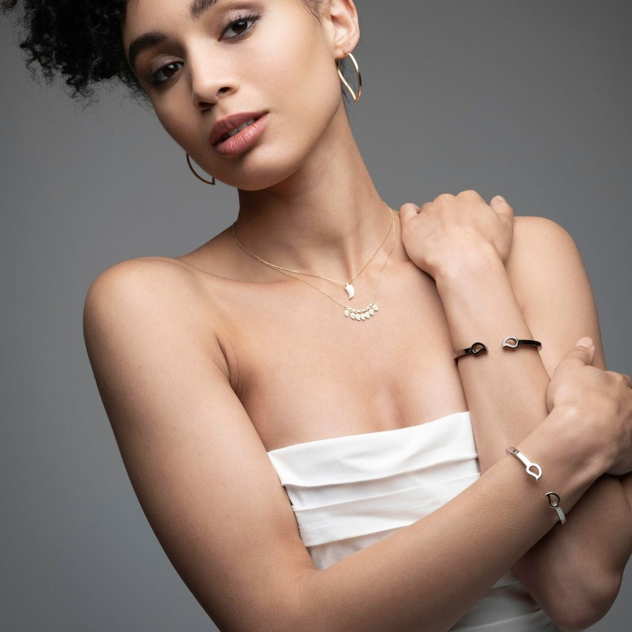 Woman wearing a silver bangle open on the front with two mango leaves on each endl, a pair of extra large hoops shaped like a mango and layered necklaces from the Inessa Love Collection